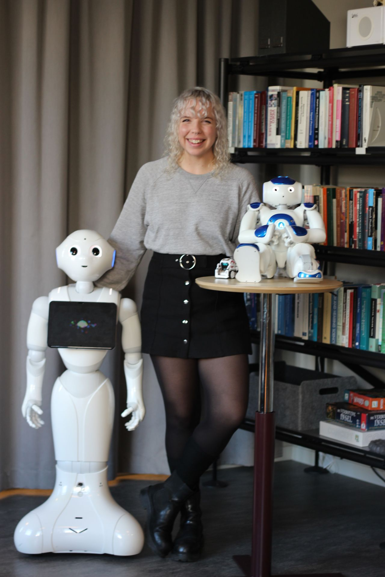 Rebecca Stower standing beside a Pepper robot, with NAO and Cozmo robots on a table